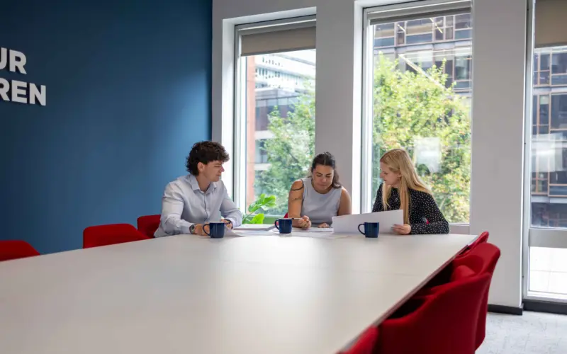 Three coworkers sit at a large conference table reviewing papers in a bright meeting room with red chairs and large windows.