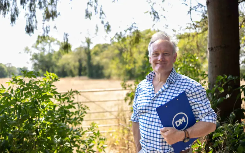 A man in a blue checkered shirt holds a blue DM folder while standing outdoors beside a tree and green shrubs, with a sunlit field behind him.