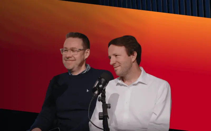 Two men stand beside a microphone in front of a red and orange gradient screen, with one wearing a navy sweater and the other a white shirt.