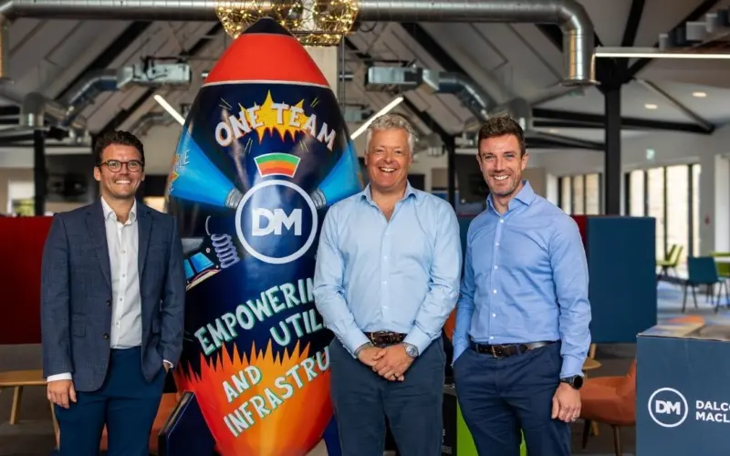 Three men in business attire standing beside a colorful rocket-shaped display with the words "One Team" and "DM" in an industrial office space.