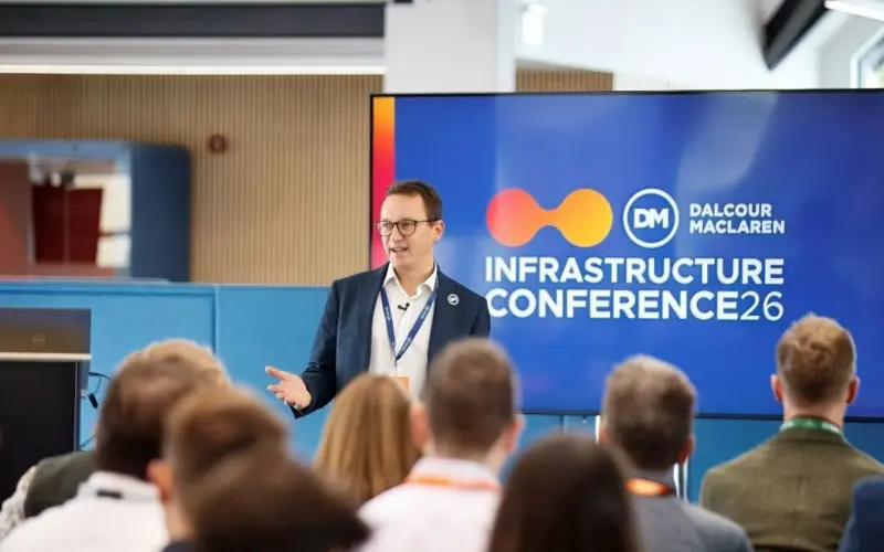 A man in a blazer speaks at the Dalcour Maclaren Infrastructure Conference 26, with an audience seated in the foreground and a presentation screen behind him.