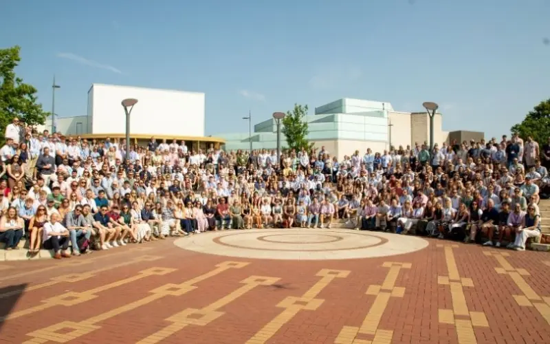 A large group of people poses outdoors on a brick plaza with a circular pattern, in front of modern campus buildings and trees.