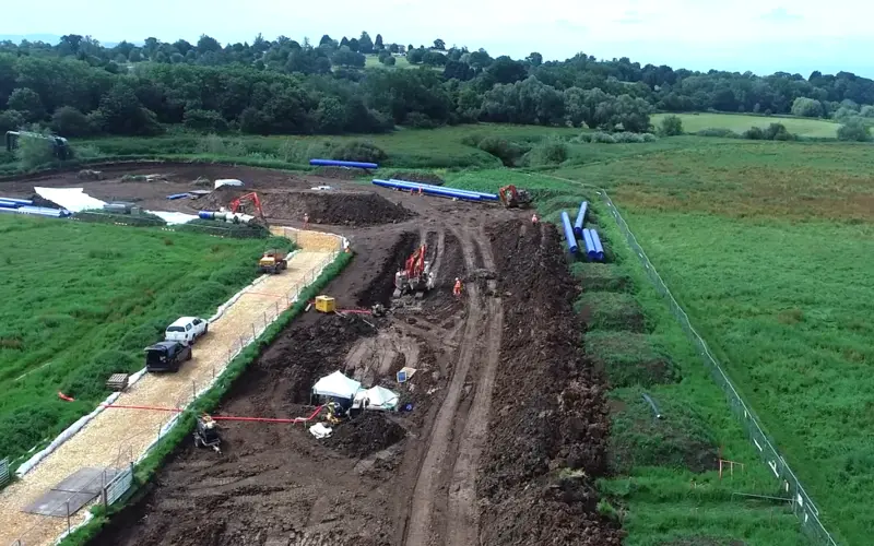 An aerial view of a construction site with heavy machinery and vehicles working on a dirt road. The area is surrounded by green fields and trees in the background under a cloudy sky.