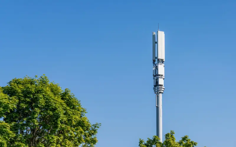 A tall cell tower with multiple antennas is visible against a clear blue sky, framed by green trees at the bottom of the image.