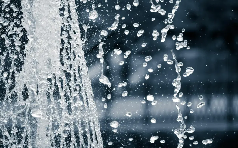 Close-up of water droplets splashing from a fountain against a blurred background. The image captures the motion and texture of the water as it cascades.