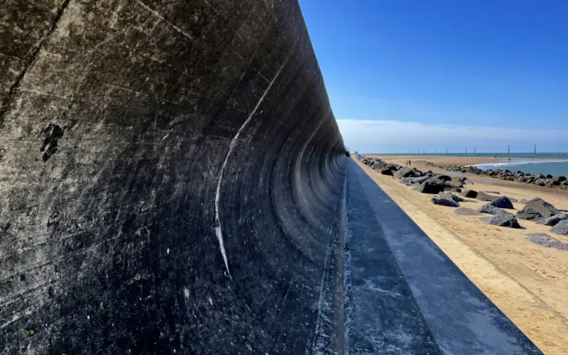 A curved concrete wall along a beach, with a coastline visible in the background. The sky is clear and blue, and a line of rocks runs along the sand beside the water.