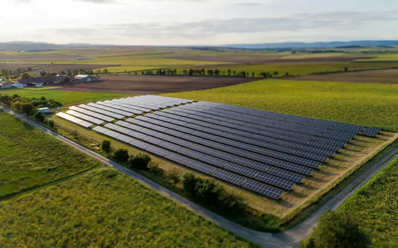 An aerial view of a solar farm with rows of solar panels arranged in a grid pattern, surrounded by green fields and farmland. A dirt road leads around the solar installation.