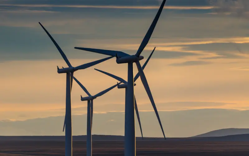 Three wind turbines silhouetted against a colorful sunset, with a vast open landscape in the background.