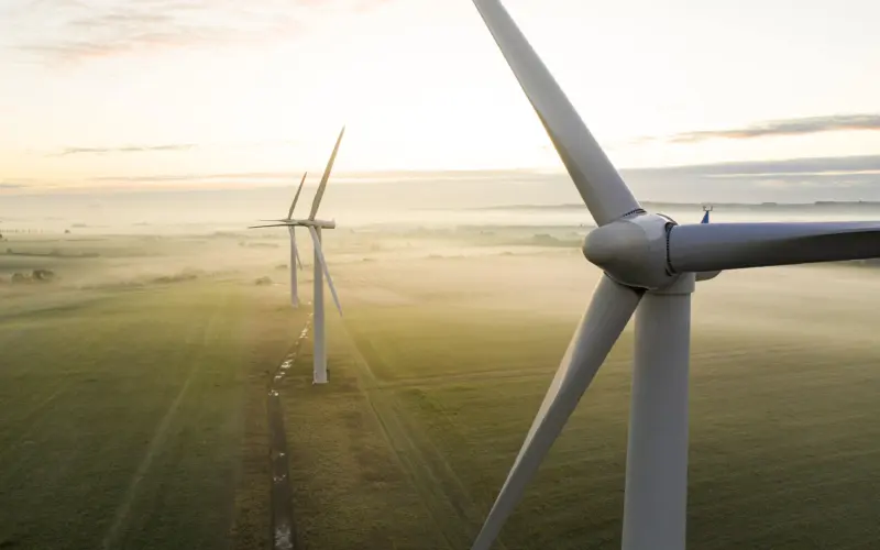 Aerial view of several wind turbines in a green landscape during early morning light, with mist covering the ground and a winding path visible in the foreground.