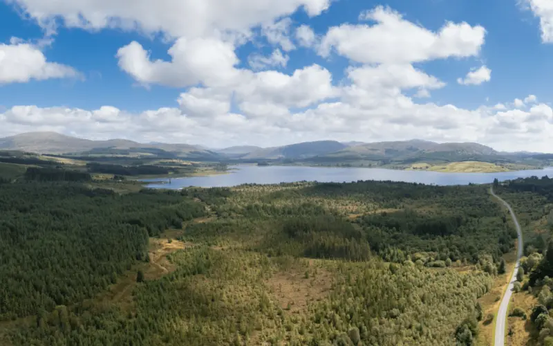 A panoramic view of a lake surrounded by green forests and rolling hills under a partly cloudy sky. A winding road is visible on the right side of the image.