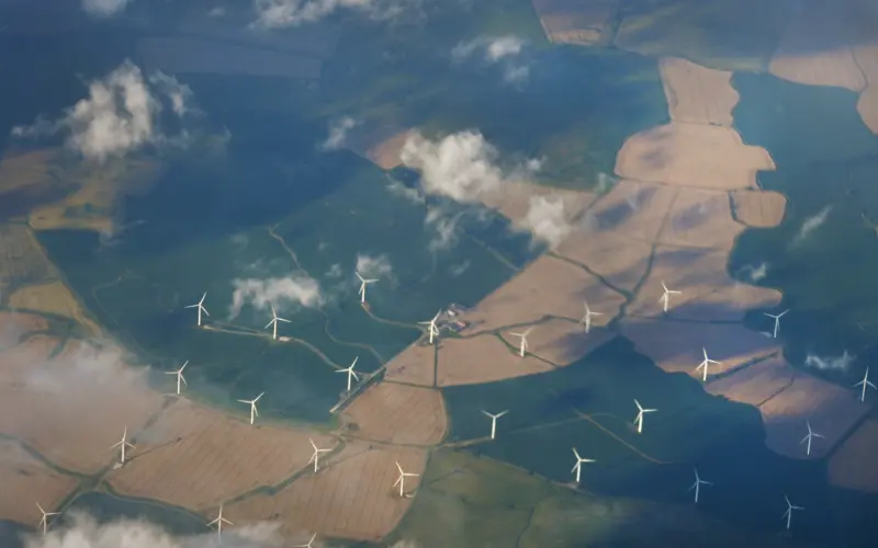 Aerial view of farmland with wind turbines scattered across the fields, under a partly cloudy sky. The landscape features patches of different colors indicating various crops.