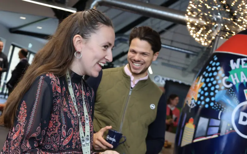 Two smiling individuals engaged in conversation at a lively event. The woman, wearing a patterned blouse, holds a small object while the man, dressed in a green vest, appears amused. A vibrant display with the text 'WE CHALLENGE' is visible in the background.