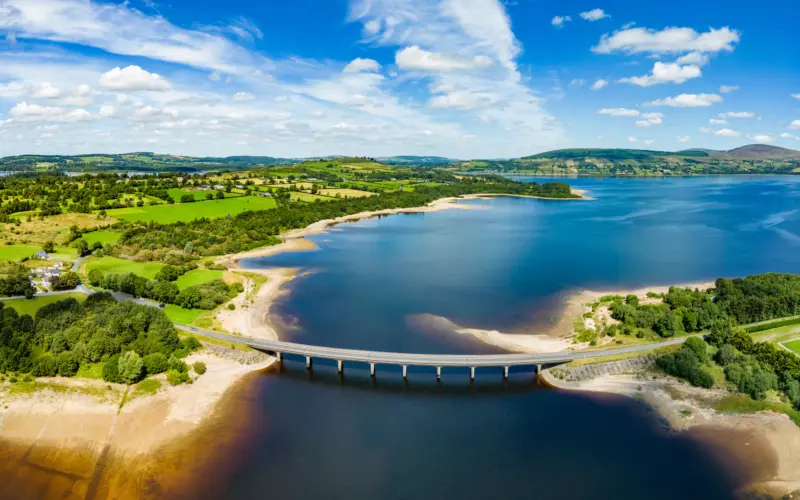Aerial view of a river with a bridge spanning across it, surrounded by lush green fields and rolling hills under a bright blue sky with fluffy clouds.