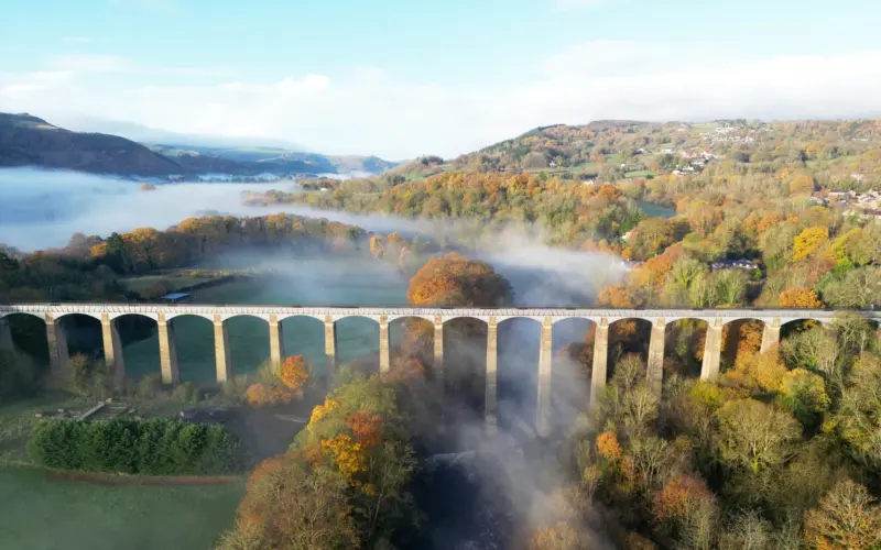 Aerial view of a stone viaduct with multiple arches spanning over a misty landscape filled with autumn-colored trees and low-lying fog.