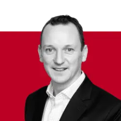 A portrait of a man in a suit with short hair, smiling, against a split background of green and red. The man is wearing a white shirt under a black blazer.