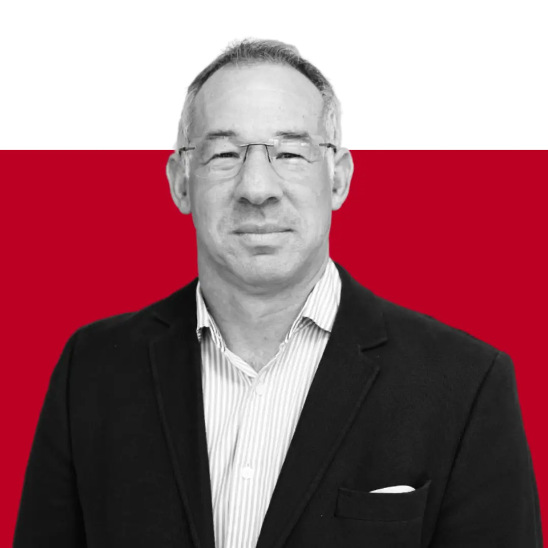 A man wearing glasses and a black blazer over a striped shirt, posing against a red background.