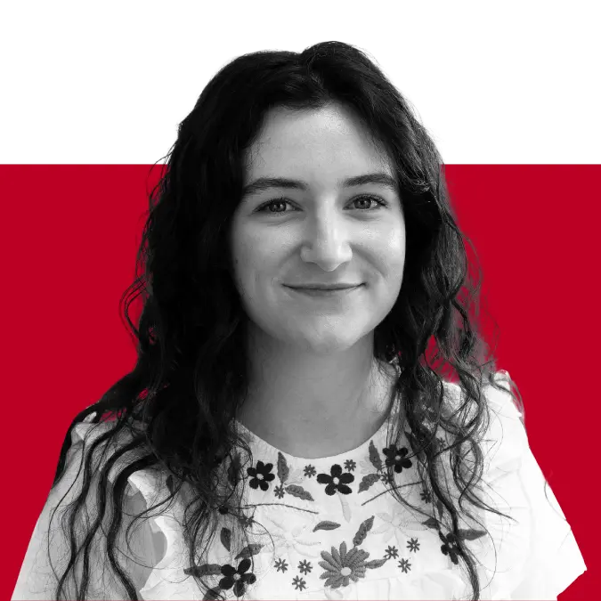 A woman with long curly hair smiles while wearing a floral patterned shirt, against a solid red and black background.