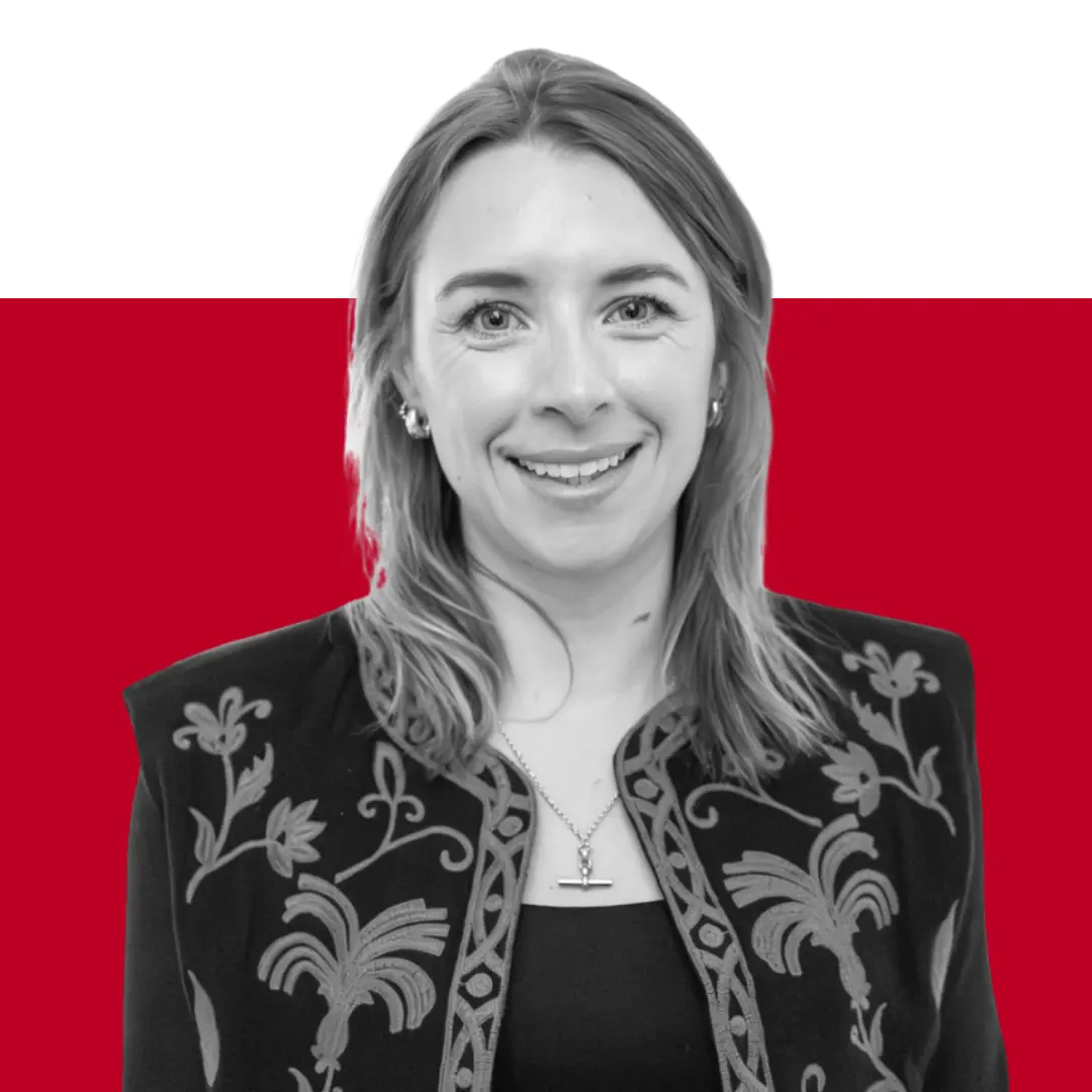 A woman with long blonde hair, wearing a black embroidered jacket, smiles for the camera. The background features a solid red and black gradient.