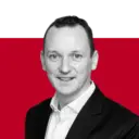 A professional headshot of a man in a black blazer and white shirt against a red background. He is smiling directly at the camera.