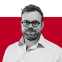A man with glasses and a beard wearing a light-colored shirt, standing against a solid red background.