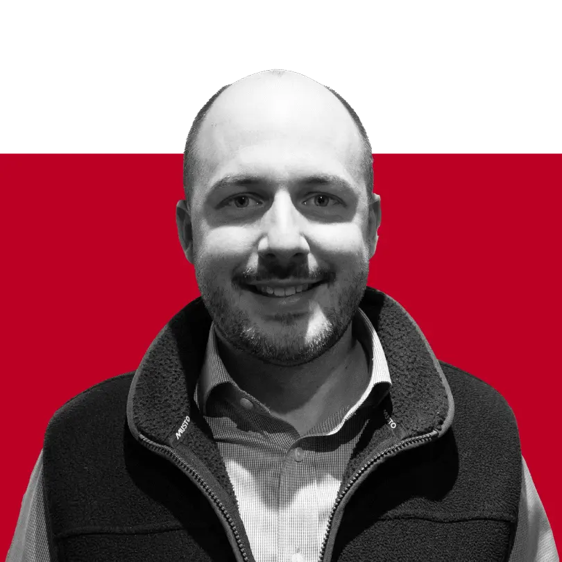 A man with a bald head and a beard, wearing a black fleece vest over a collared shirt, smiles in front of a solid red background.