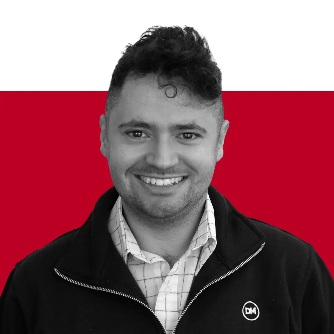 A man with dark, curly hair smiling at the camera, wearing a black jacket over a checked shirt, set against a red and black background.