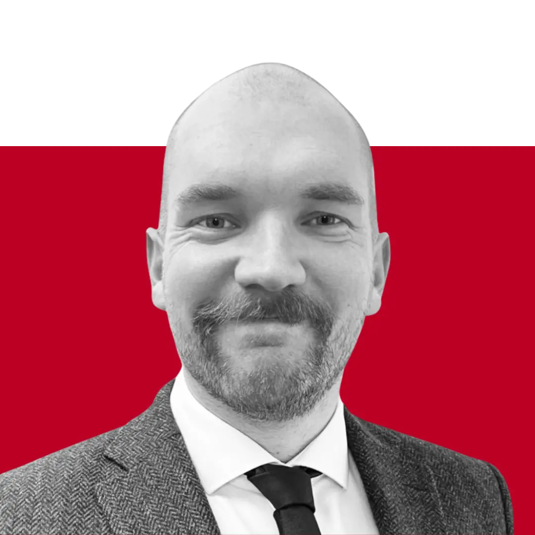 A man with a shaved head and a beard is smiling while wearing a gray suit and a white shirt with a black tie. The background is a solid color, transitioning from black to red.