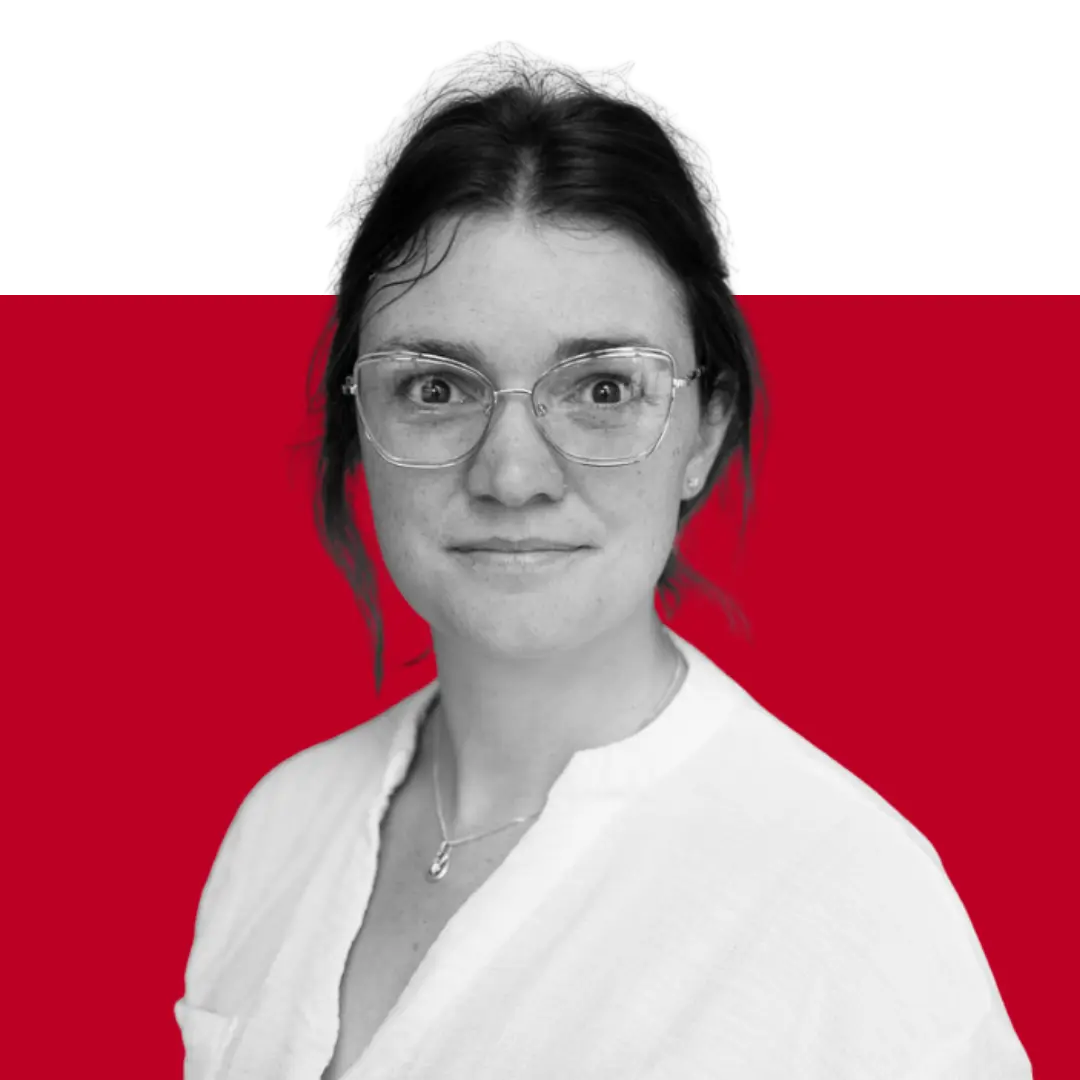A woman with glasses and dark hair is positioned against a red background, looking directly at the camera. She wears a white blouse and has an attentive expression.