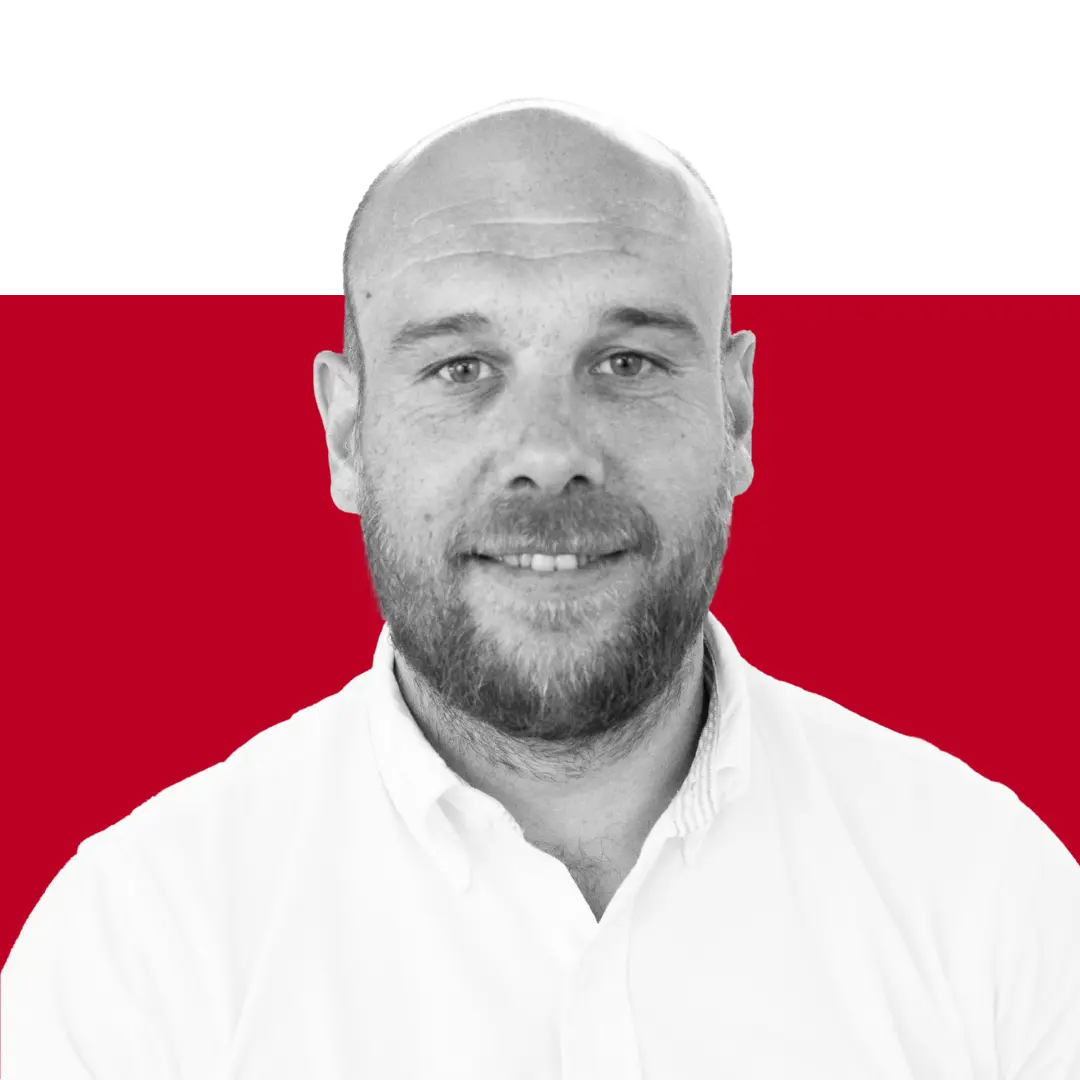 A smiling man with a shaved head and a beard, wearing a white collared shirt, is photographed against a red and black background.