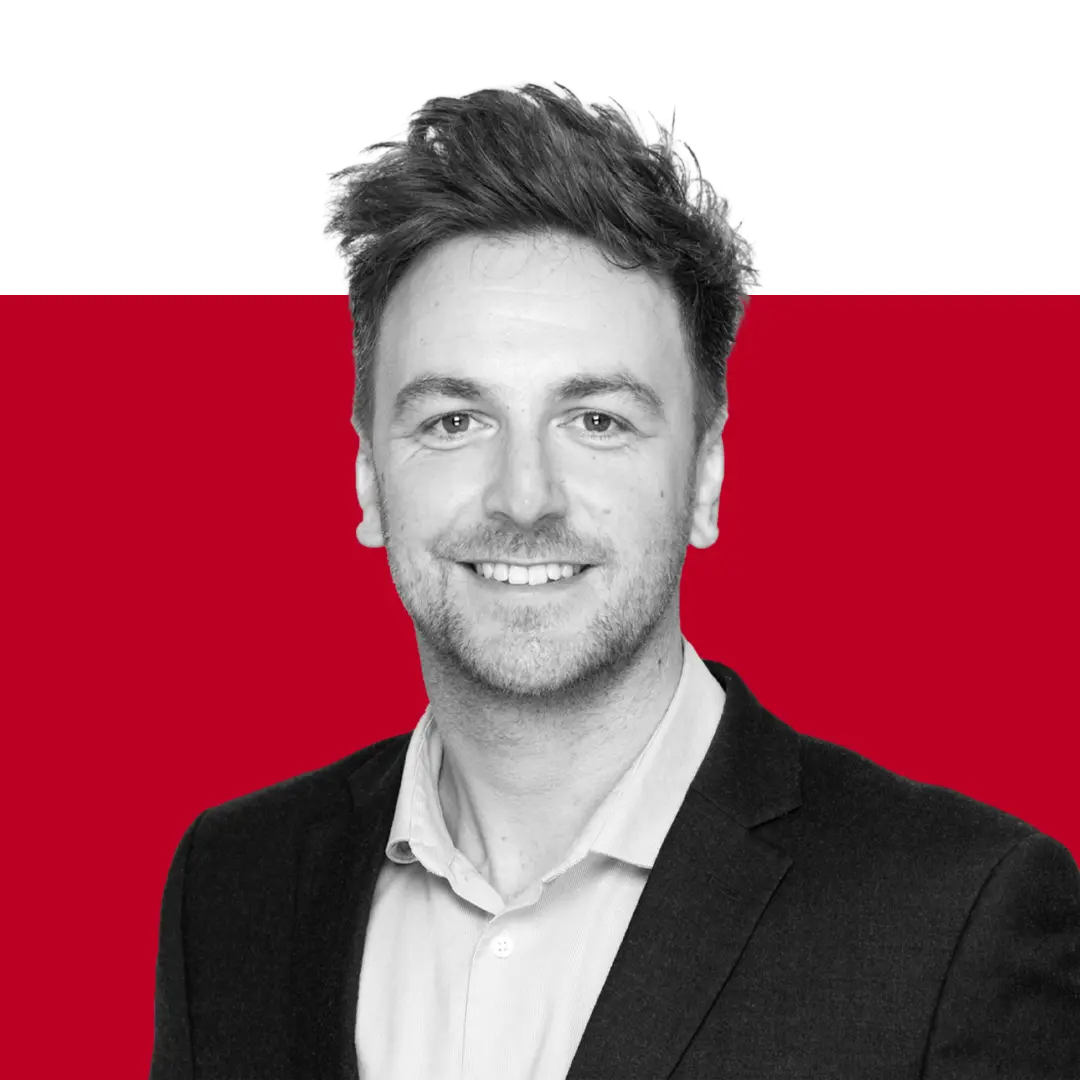 A professional headshot of a man smiling, wearing a black suit and a light-colored shirt, with a red and black background.