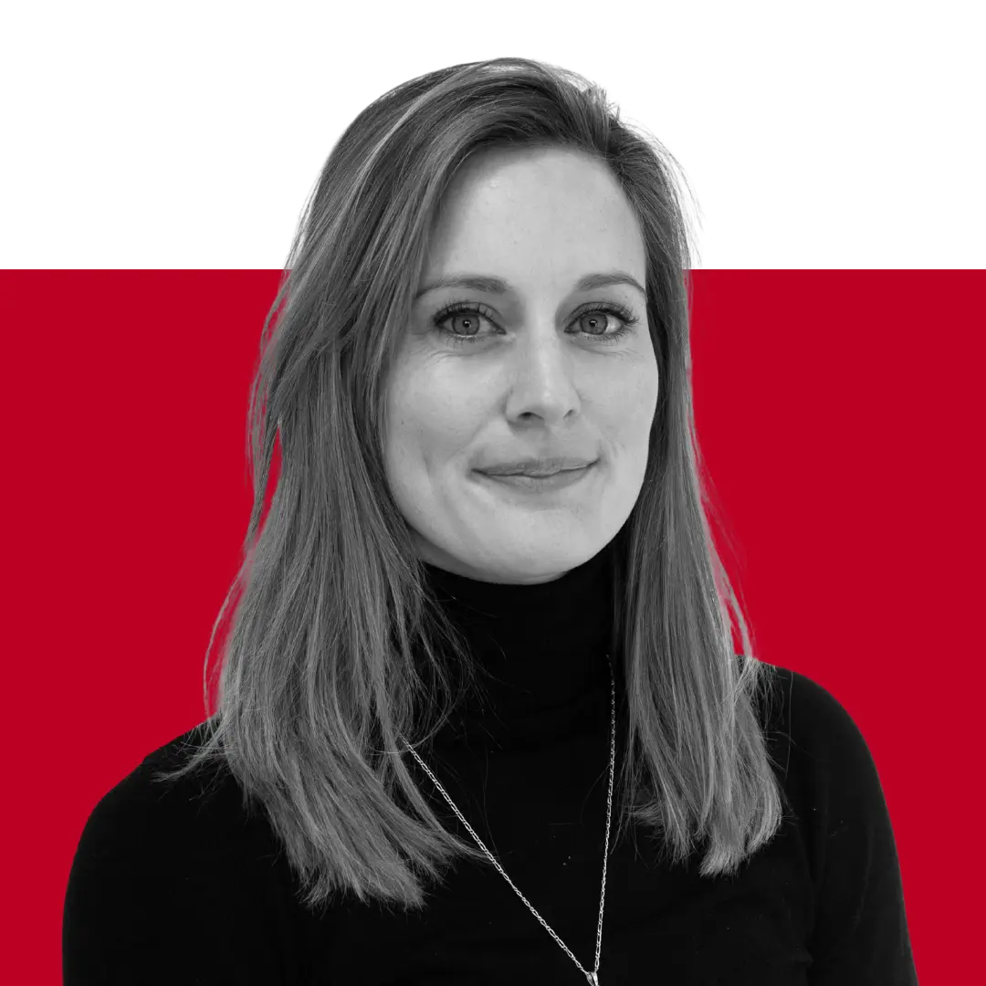 A woman with long brown hair wearing a black turtleneck sweater, smiling while facing the camera. The background features a solid black upper section and a solid red lower section.
