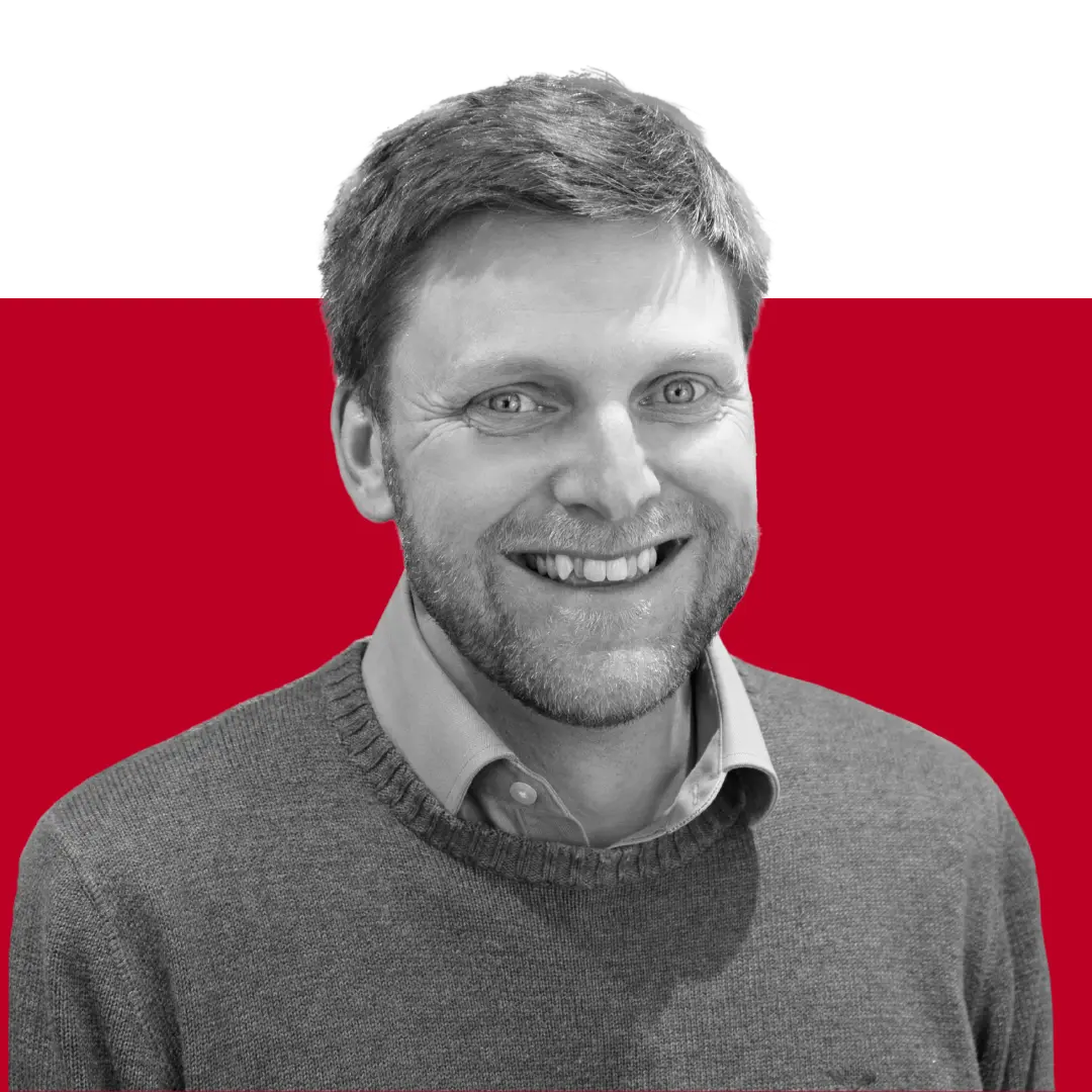 A smiling man with short hair and a beard, wearing a grey sweater over a collared shirt, against a red and black background.