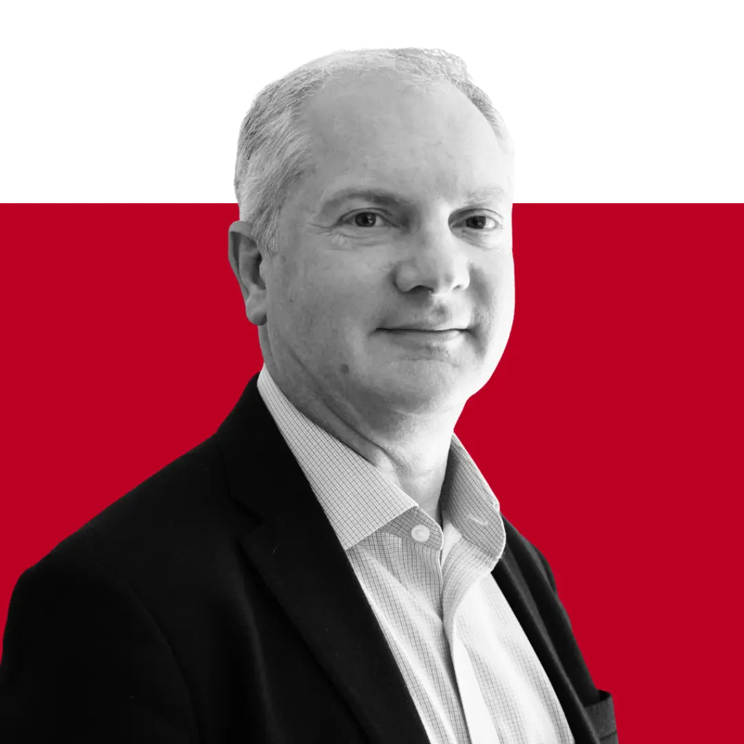 A man with short, light-colored hair and wearing a black blazer over a collared shirt, posing against a red background. He is looking slightly towards the camera.