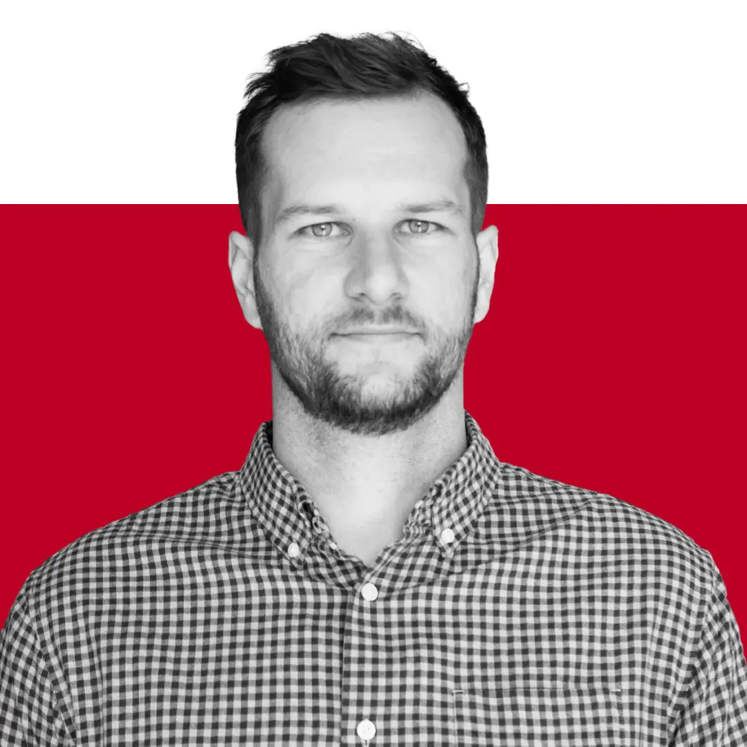 A man with short hair and a beard, wearing a checkered shirt, stands against a solid red background, looking directly at the camera.