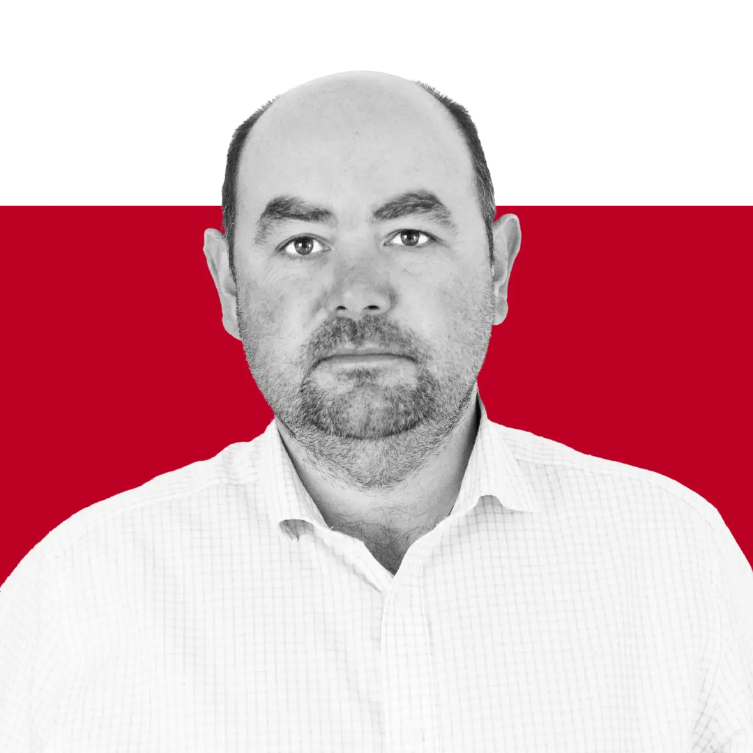 A man with a beard wearing a white button-up shirt and posing against a red and black background. He has a neutral expression and is looking directly at the camera.
