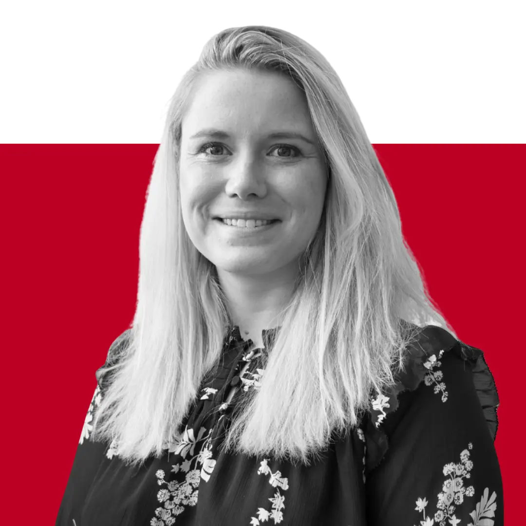 A woman with long blonde hair, wearing a floral black blouse, smiles at the camera. The background features a solid red color.