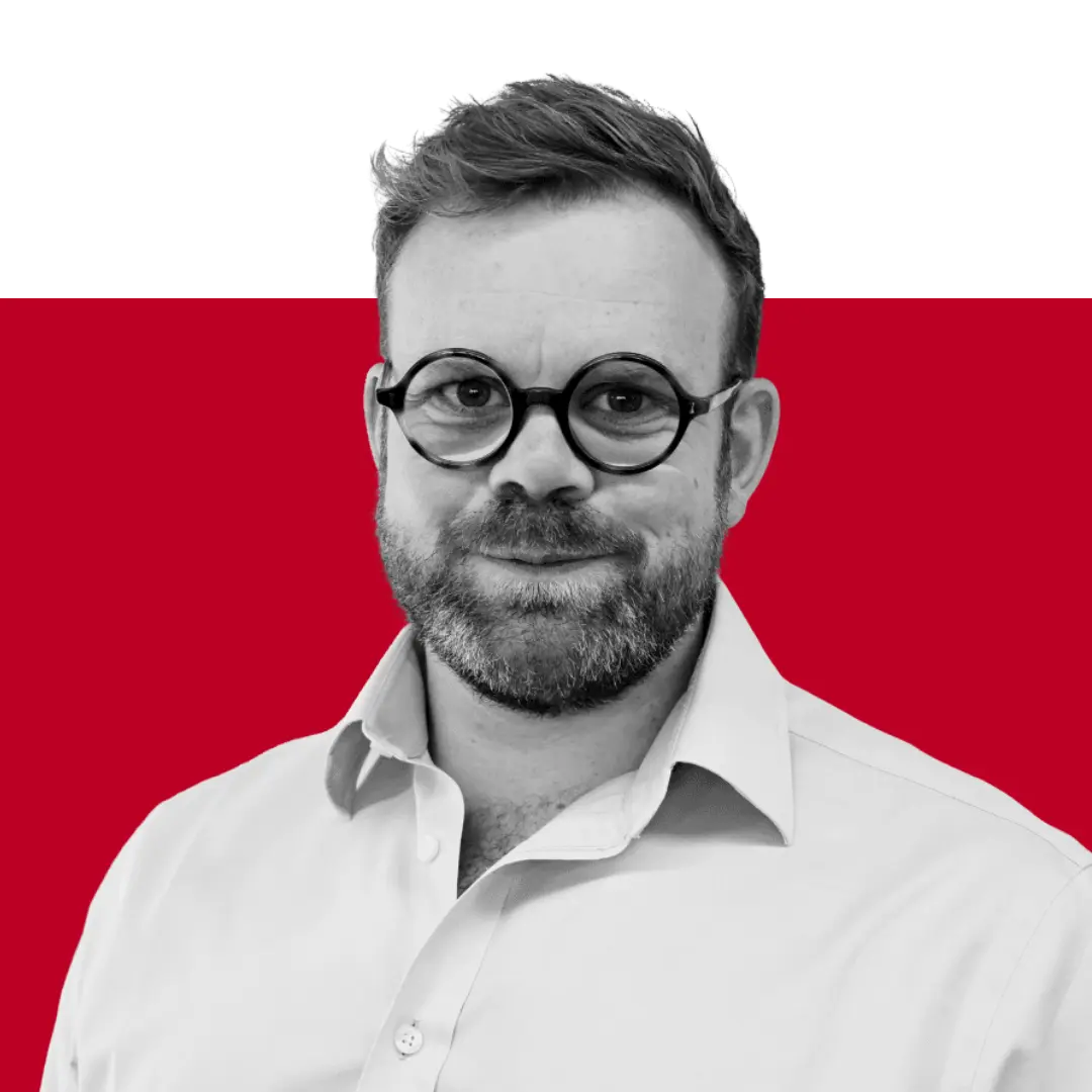A man with glasses and a beard wearing a light-colored shirt, standing against a solid red background.