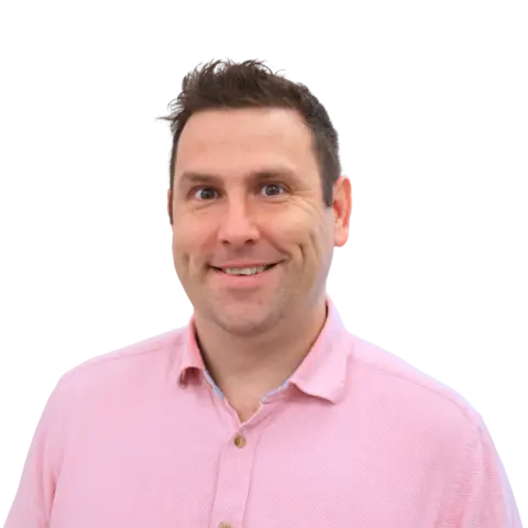 A man smiling and looking at the camera, wearing a light pink collared shirt. The background is plain white, creating a clean and simple portrait.