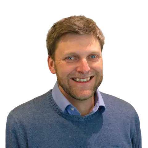 A man with short brown hair and a beard, wearing a blue sweater over a collared shirt, smiling at the camera against a plain background.