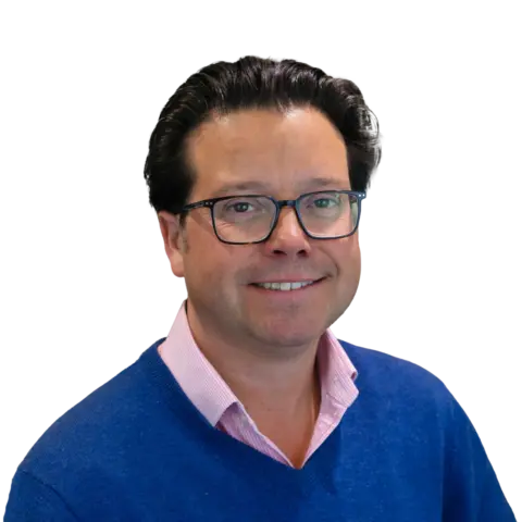 A man wearing glasses and a blue sweater over a pink collared shirt, smiling against a white background.
