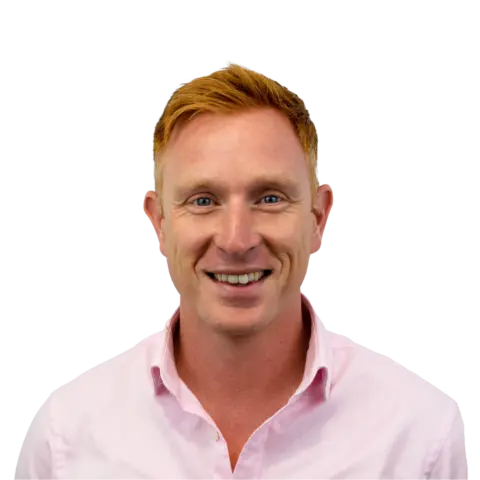 A man with short red hair smiling at the camera, wearing a light pink button-up shirt, against a plain white background.