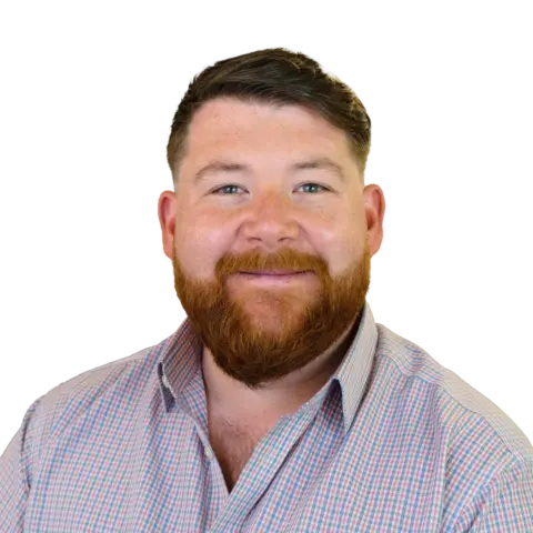 A man with a beard, wearing a plaid button-up shirt, smiles at the camera. The background is plain, emphasizing his facial features and expression.