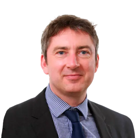A man wearing a dark suit and a checked shirt, with a tie, smiling against a plain background.