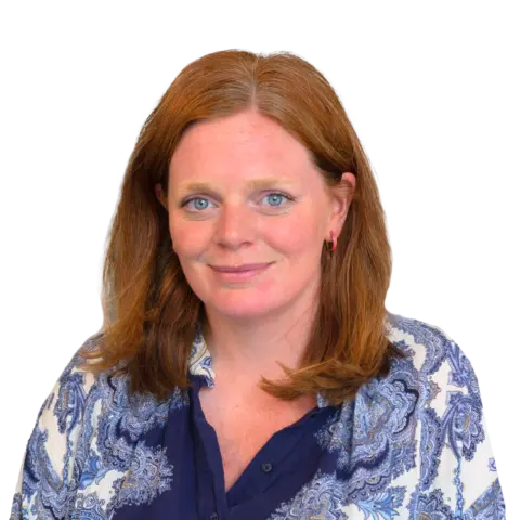 A woman with long, wavy reddish-brown hair, wearing a blue and white patterned blouse, smiles in front of a plain background. She has blue eyes and is partially facing the camera.