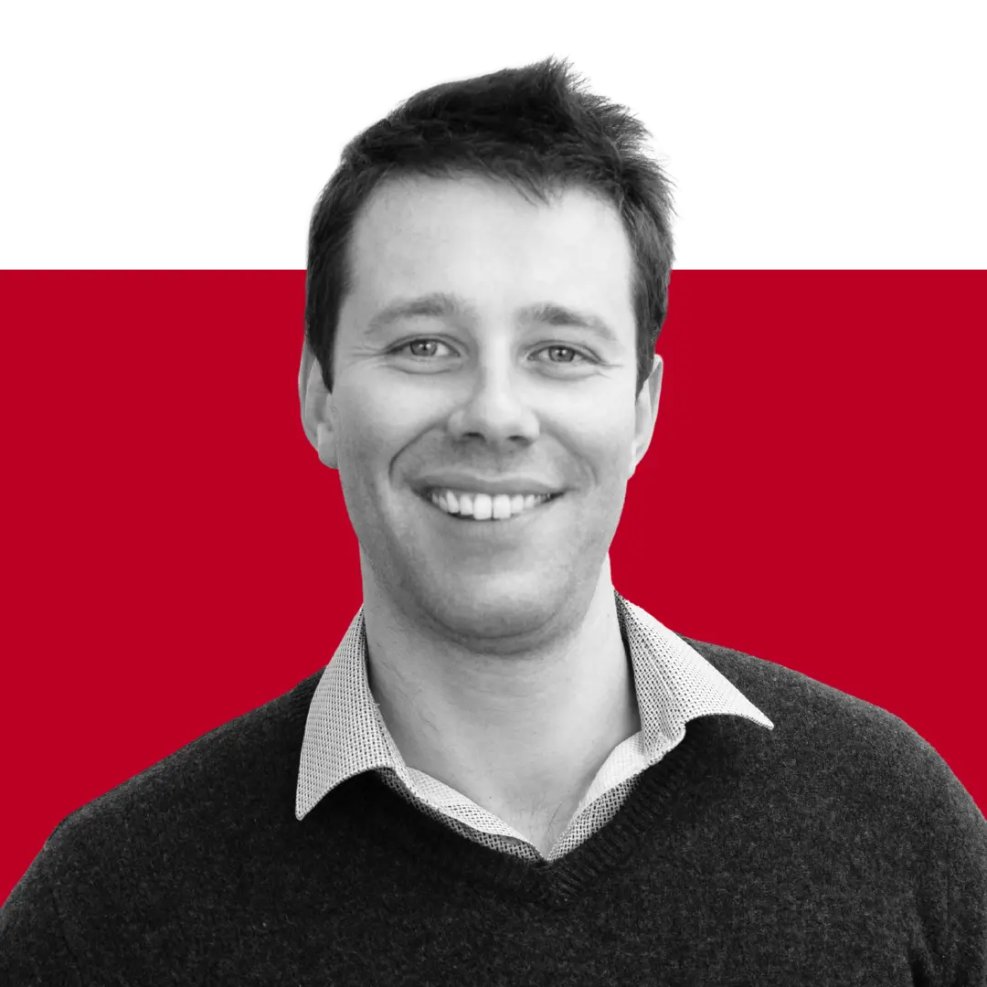 A smiling man with short dark hair, wearing a light gray collared shirt and black sweater, set against a black and red background.