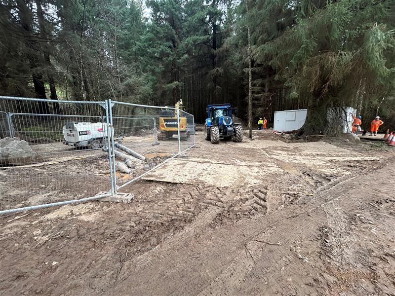 A construction site in a wooded area, featuring construction machinery including a blue tractor and yellow excavator, surrounded by a temporary fence. Work vehicles and workers in safety gear are visible among trees.