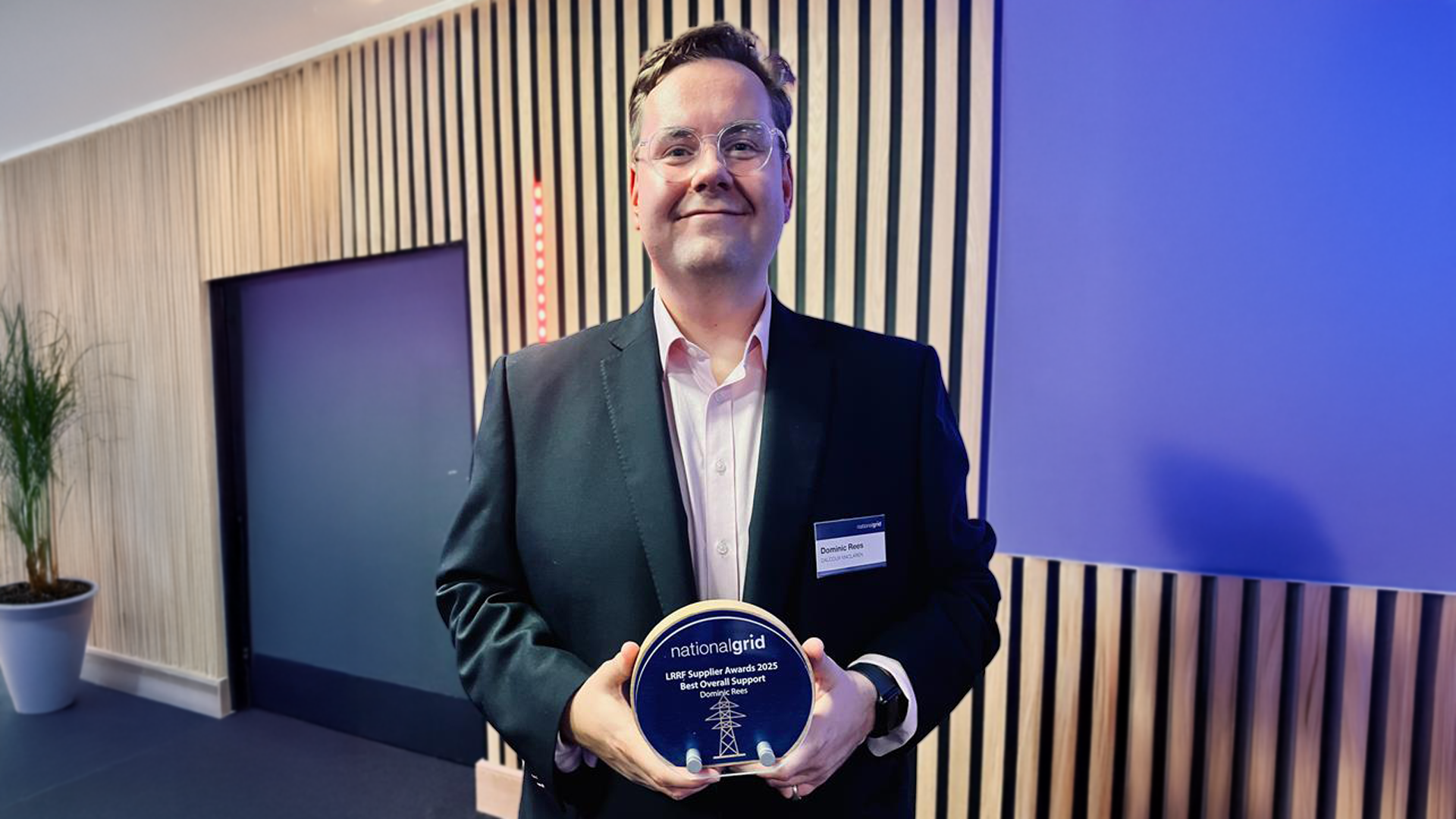 A man standing in a modern room, holding an award plaque with the text "National Grid Award". He is wearing a black suit and a white shirt, with a name tag on his jacket. The background features wooden paneling and potted plants.