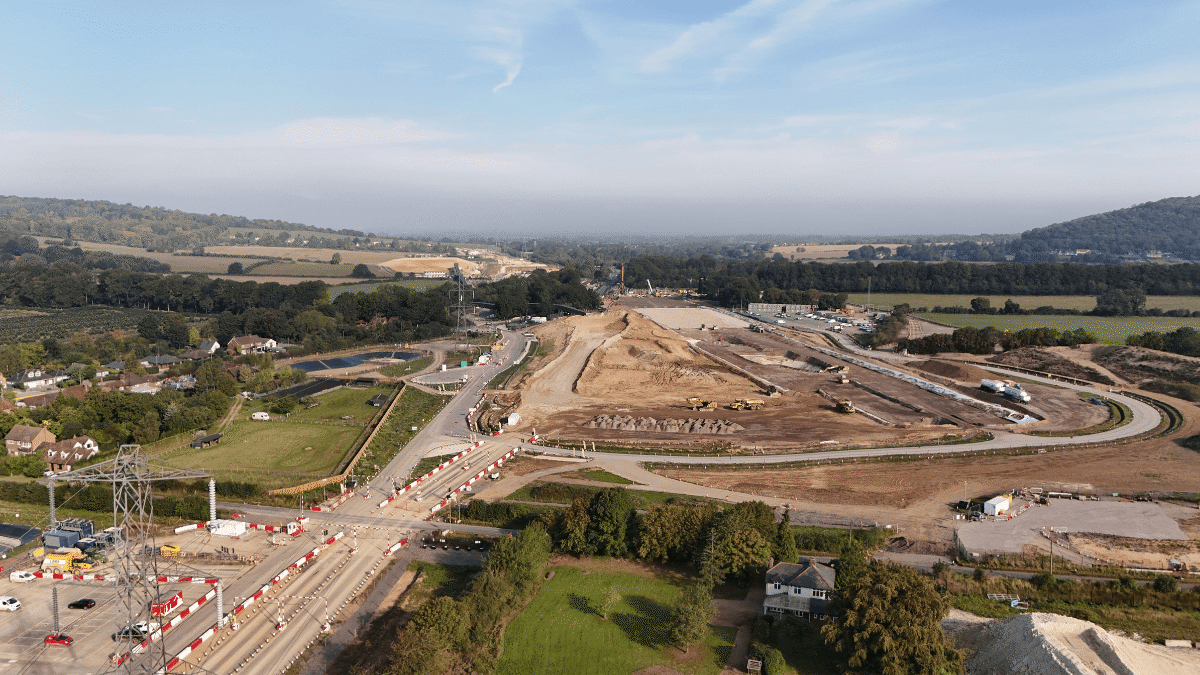 Aerial view of a large construction site surrounded by hills and farmland. The area shows piles of earth and construction equipment, with a road leading to the site and traffic cones marking the area.