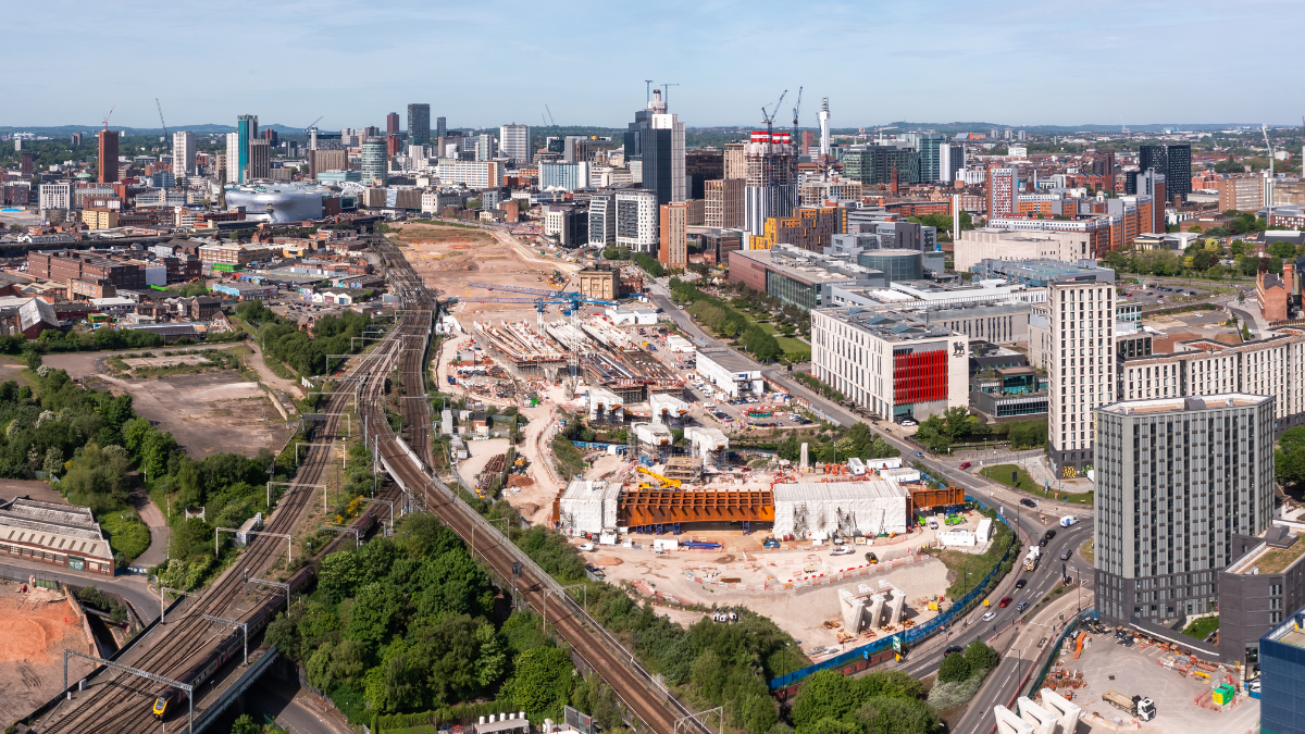 Aerial view of a city skyline with various buildings and cranes, showcasing a construction site surrounded by greenery and rail tracks. The scene features urban development under a clear blue sky.