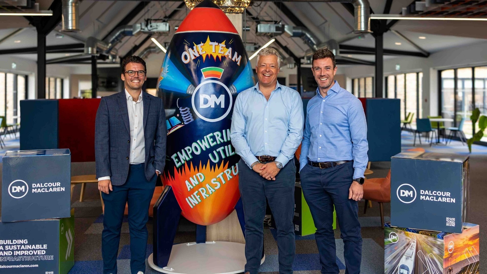 Three men in business attire stand beside a colorful surfboard-shaped display reading “One Team DM” and “Empowering Communities,” inside a modern office space with Dalcour Maclaren signs.
